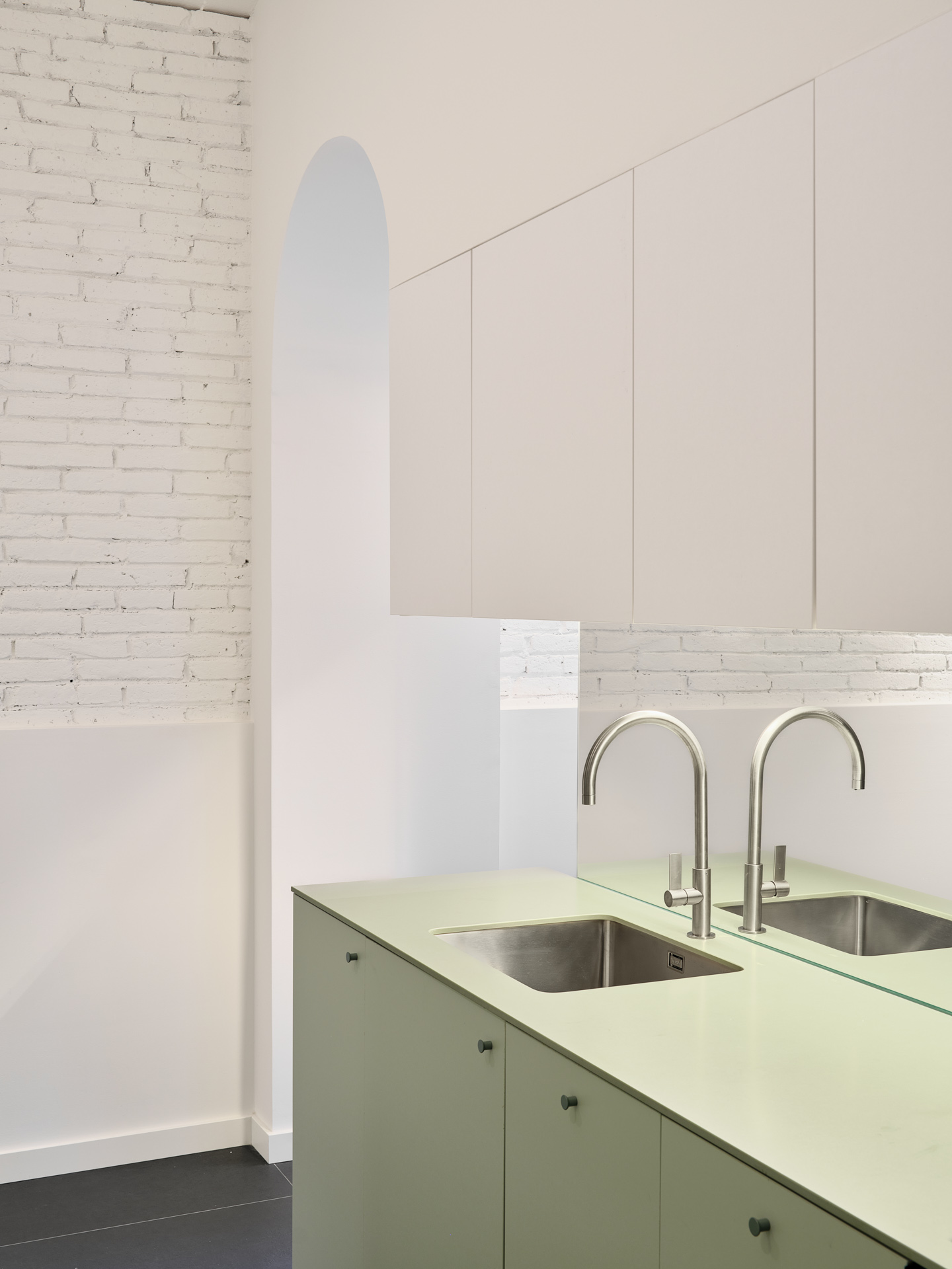 Kitchen detail in CRÜ’s “La Dolors” renovation, with a pale green island featuring stainless-steel sinks, a mirrored backsplash, and whitewashed brick walls.